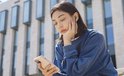 Vrouw met zwarte oordopjes en blauwe trui, denkend met telefoon in de hand.