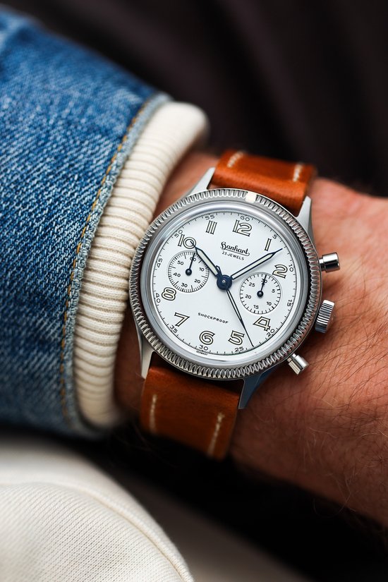 Zilverkleurig horloge met witte wijzerplaat, bruine leren band, gedragen op een pols met spijkerjack.