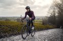 Fietser op kasseien draagt een bordeauxrood wielershirt met lange mouwen en zwarte fietsbroek. Glanzende bril op het hoofd.
