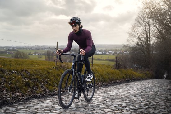Fietser op kasseien draagt een bordeauxrood wielershirt met lange mouwen en zwarte fietsbroek. Glanzende bril op het hoofd.