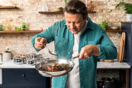 Jamie Oliver lepelend jus over vlees in een RVS koekenpan.