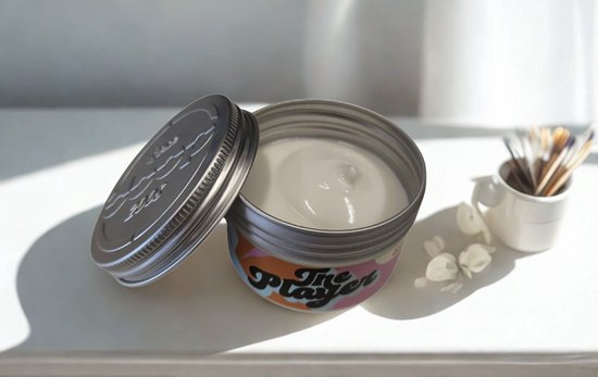Een open aluminium potje met witte crème. De pot heeft een kleurrijk label met de tekst 'The Player'.