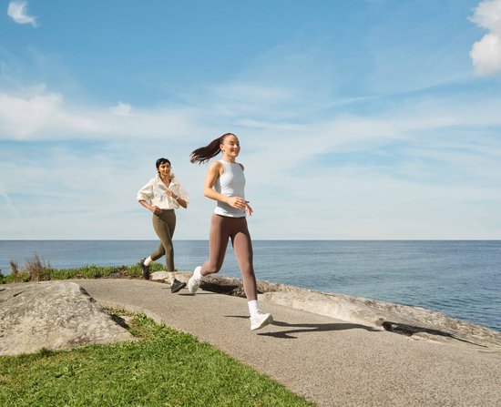 Twee vrouwen rennen langs de kustlijn met op de voorgrond een vrouw in een blauw sport-topje en bruine legging.
