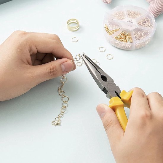 Twee handen gebruiken een tang om gouden ringetjes te verbinden voor een armband, met een rond doosje met ringetjes ernaast.