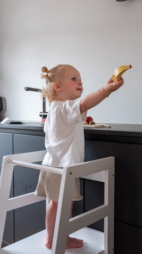 Kind met blonde knotjes in wit shirt en beige broek op witte leertoren, houdt houten banaan omhoog.