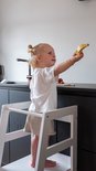 Kind met blonde knotjes in wit shirt en beige broek op witte leertoren, houdt houten banaan omhoog.