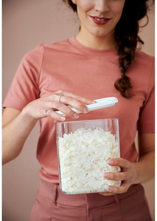 Une femme avec une tresse tient une boîte de conservation transparente avec couvercle blanc, remplie de flocons de noix de coco.