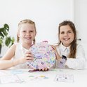 Deux filles souriantes assises à une table, l'une tenant un coffret créatif en forme de cœur sur le thème de la licorne.