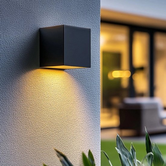 Zwarte kubusvormige buitenlamp aan een grijze muur, straalt warm licht naar beneden.