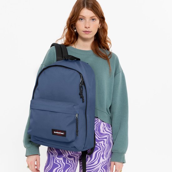 Femme aux cheveux roux porte un sac à dos Eastpak bleu marine sur son épaule, avec un pantalon à rayures violettes.