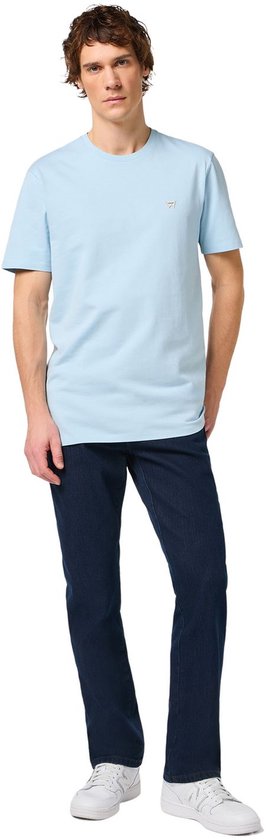Model draagt een lichtblauw T-shirt en een donkerblauwe Wrangler regular fit jeans met witte sneakers.