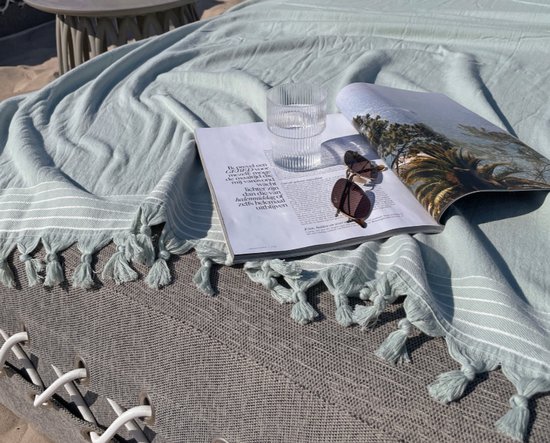 Jadegroene hamamdoek met witte strepen en kwastjes, liggend op zand, naast een tijdschrift en zonnebril.