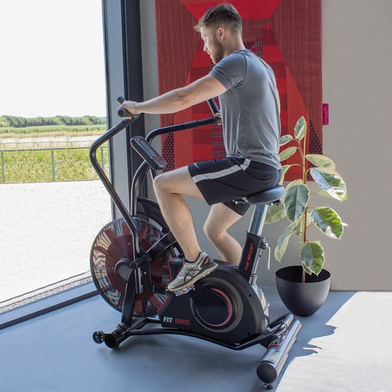 Man traint op een zwart 'FIT BIKE The Beast' airbike met rood ventilatorwiel, staand naast een grote plant.