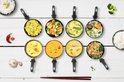 Bovenaanzicht van 8 wokpannetjes gevuld met diverse gerechten: pasta, groenten, curry, garnalen en noedels.