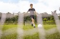 Jongen traint voetbal op grasveld met voetbaldoel van AJ-Sports op de achtergrond.