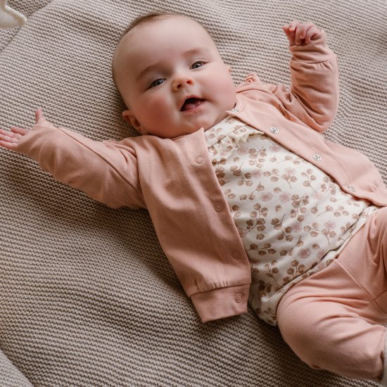 Baby met grote blauwe ogen ligt op een beige plaid en draagt een roze vestje over een wit jurkje met bloemenprint.