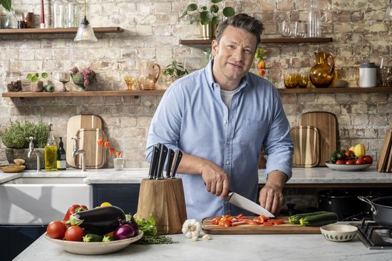 Jamie Oliver in blauw hemd snijdt rode pepers op een acaciahouten snijplank in een keuken.
