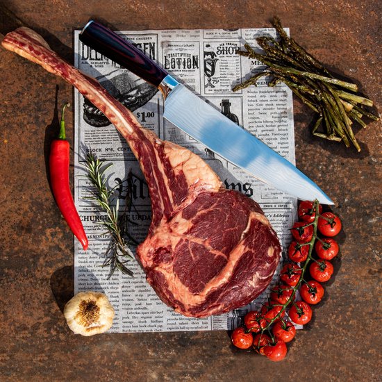 Une grosse côte de bœuf avec un couteau, du piment, de l'ail, de l'aneth, des tomates et des asperges sur un journal.