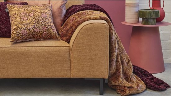 Gouden plaid met een bruin-roze bloemenprint, gedrapeerd over een okerkleurige bank.