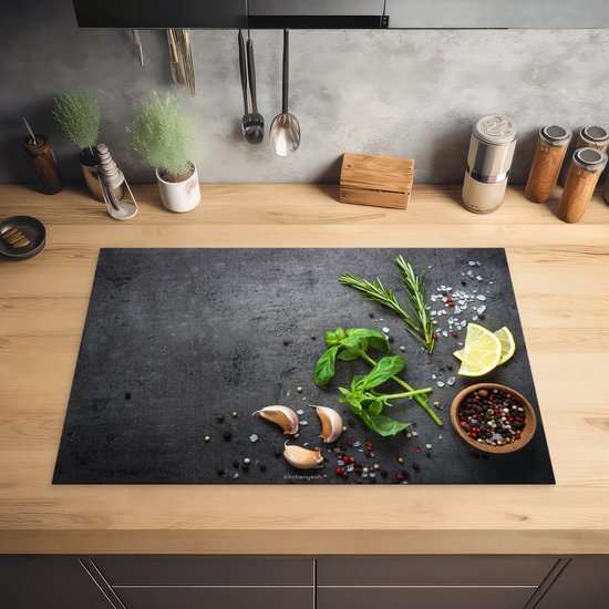 Zwarte inductie beschermer met betonprint, basilicum, rozemarijn, citroenpartjes, knoflook en peperkorrels op een houten aanrecht.