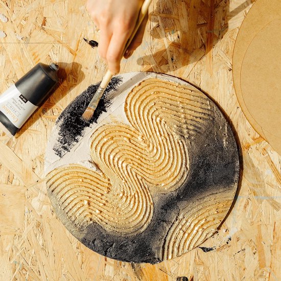 Een hand met penseel brengt gouden textuurpasta aan op een rond kunstwerk met zwarte achtergrond.