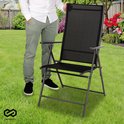 Man in wit hemd en grijze broek staat naast een zwarte inklapbare tuinstoel in een tuin.