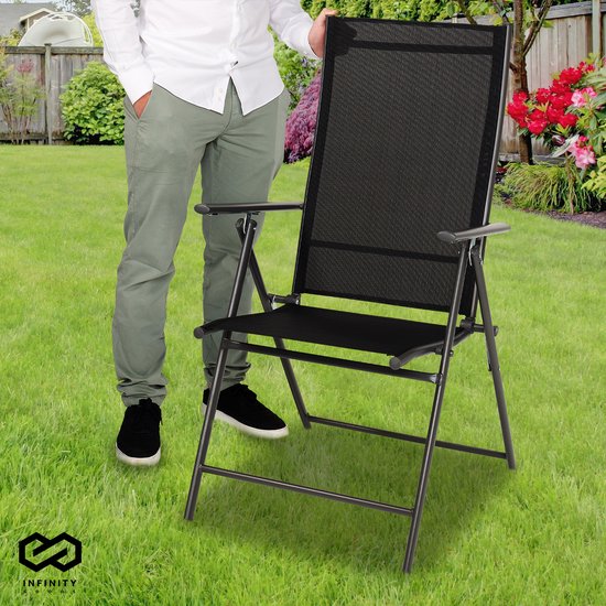 Man in wit hemd en grijze broek staat naast een zwarte inklapbare tuinstoel in een tuin.