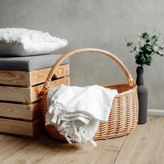 Panier en osier avec une couverture blanche à franges, à côté de caisses en bois et d'un vase avec des plantes vertes.