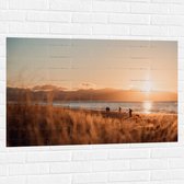 WallClassics - Muursticker - Zon Verdwijnend achter Bergen bij Strand - 105x70 cm Foto op Muursticker