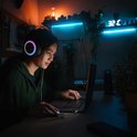 Jonge vrouw met gaming koptelefoon met led-verlichting, kijkt naar laptop in kamer met blauwe en oranje led-verlichting.