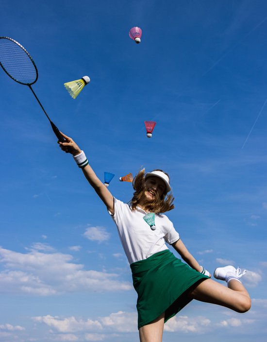 Meisje speelt badminton, gooit 5 kleurrijke shuttles de lucht in. Ze draagt witte sportkleding en groene rok.