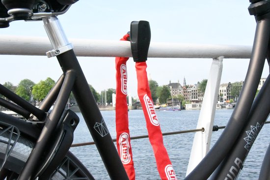 Fietsketting ABUS rood, bevestigd aan een fietsframe bij een waterkant met huizen op de achtergrond.