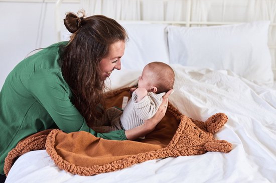 Vrolijke vrouw knuffelt met een baby op een bed, bedekt met een bruine deken met berenoortjes.