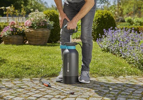 Persoon met grijze jeans en grijze sneakers drukt op de pomp van een donkergrijze 5L drukspuit met turquoise accenten.