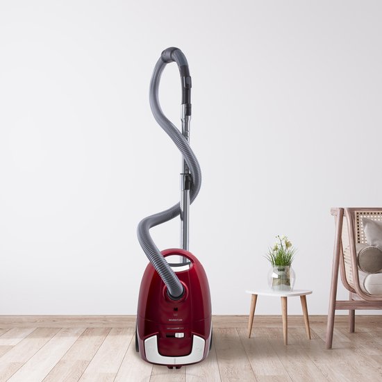 Rode stofzuiger met grijze slang en zilveren buis, staand op een houten vloer naast een bijzettafel met plant.