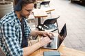 Man met blauwe geruite blouse draagt een donkerblauwe koptelefoon met geometrisch patroon en werkt op een laptop in een café.