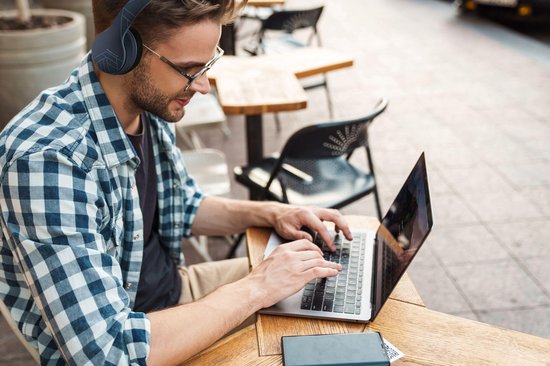 Man met blauwe geruite blouse draagt een donkerblauwe koptelefoon met geometrisch patroon en werkt op een laptop in een café.