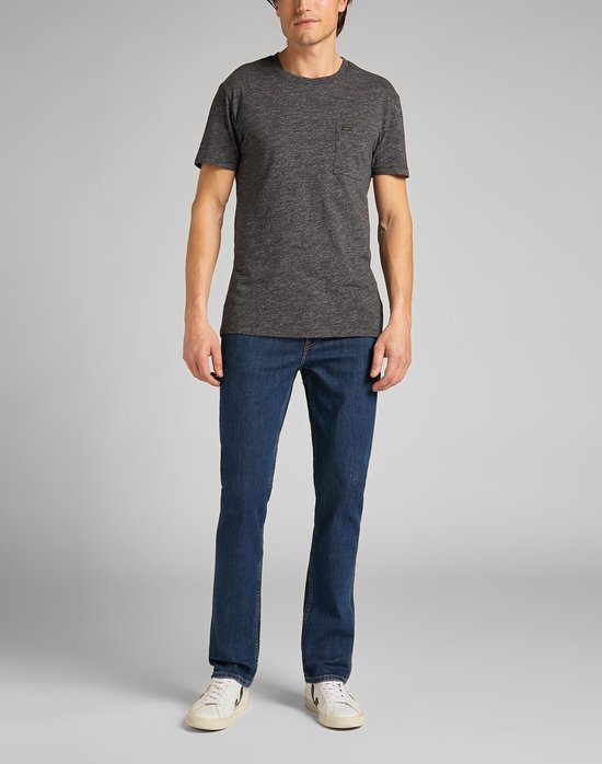 Man in een blauwe jeans met rechte pijpen, grijs T-shirt en witte sneakers, staand tegen een grijze achtergrond.