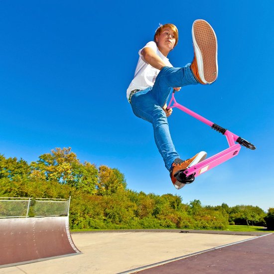 Jongen met roze stuntstep maakt een sprong in de lucht bij een skatepark. Hij draagt een witte T-shirt en blauwe jeans.