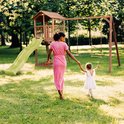 Moeder in roze jurk wandelt met dochter bij speeltoestel in park.
