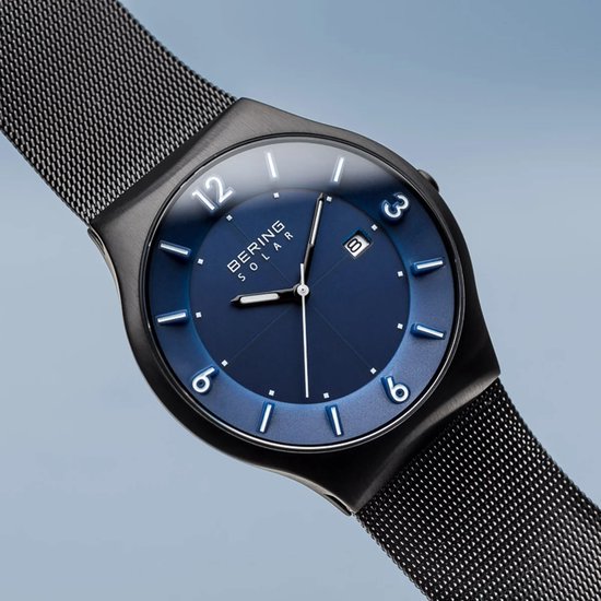 Close-up van een zwart horloge met blauwe wijzerplaat, zilveren indexen en datum. Milanese band.