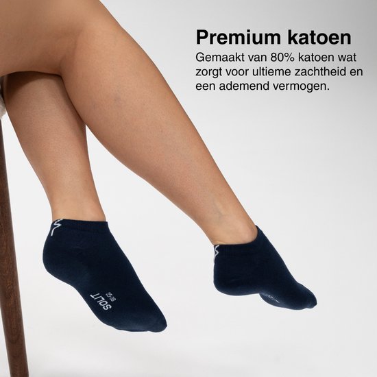 Zittende persoon met benen gekruist, draagt donkerblauwe enkelsokken met witte logo's en een donkerbruine stoel.