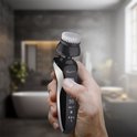 Hand houdt zwarte elektrische gezichtsborstel in een badkamer met een bad en spiegel op de achtergrond.