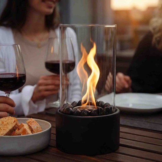 Tafelhaard met vlam, glazen cilinder, zwarte basis met stenen, op een tafel met glazen rode wijn en toastjes.