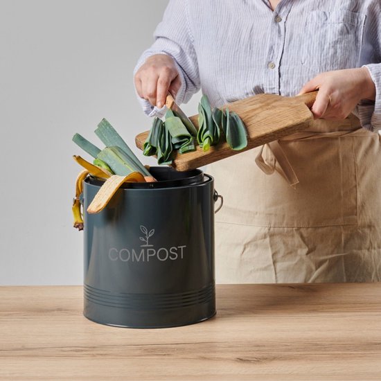 Hand voegt gesneden groenten toe aan een donkergrijze compostbak met houten handvat.