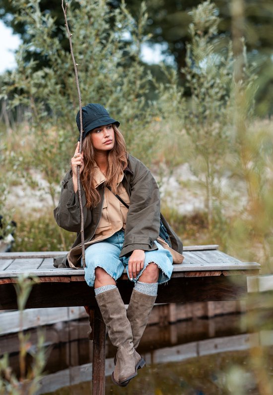 Model in de natuur, zittend op een houten bankje, draagt taupe suède laarzen, een geruite rok, spijkerbroek en een donkere hoed.