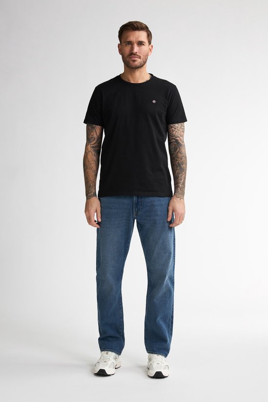 Man met tatoeages draagt een zwarte T-shirt en blauwe spijkerbroek, vooraanzicht.