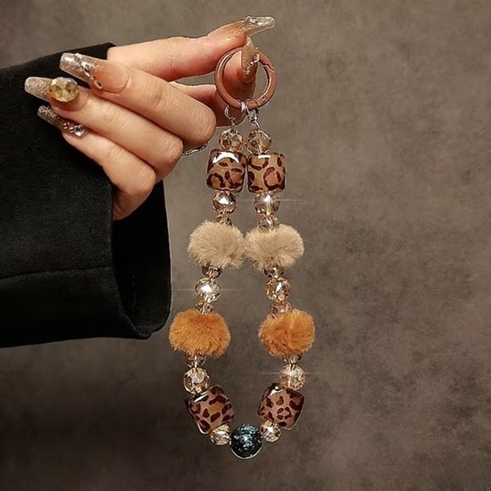 Hand met lange nagels houdt telefoonketting met bruine pompons, luipaardprint kralen en kristallen.