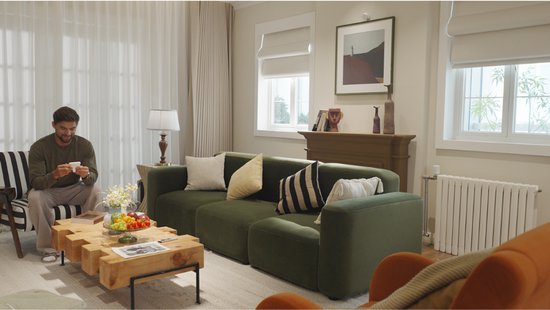 Man zit op een groene bank in een woonkamer, een witte radiator met thermostaat staat rechts.