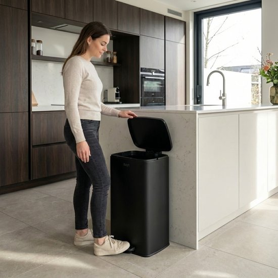 Vrouw met beige trui en zwarte jeans gebruikt een zwarte Bayt prullenbak in een moderne keuken, met het deksel open.